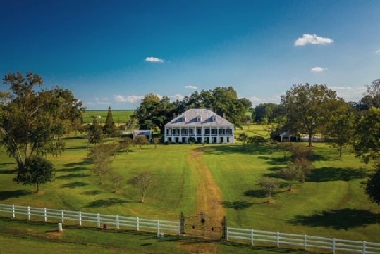 Large white plantation-style house surrounded by expansive green lawns, trees, and a white fence under a blue sky with scattered clouds.