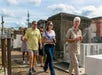 A group of people tours an outdoor cemetery, led by an older woman in a light outfit. They walk past stone tombs under a partially cloudy sky.