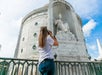 A person takes a photo of a large marble statue seated in a niche, with "Italia" inscribed below. A partially cloudy sky is visible in the background.
