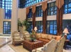 Hotel lobby with large windows, elegant seating area with a sofa, armchairs, and a wooden table, surrounded by potted plants and lamps.