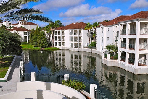 White apartment buildings with red roofs surround a calm canal. Palm trees and lush greenery line the water's edge under a partly cloudy sky.
