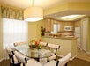 A dining room with a glass table, six chairs, and a pendant light. The kitchen with wooden cabinets and white appliances is visible in the background. A floral centerpiece is on the table.