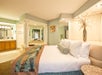 Hotel room with a large bed, decorative pillows, a patterned throw, and a vase with flowers. Open bathroom area with a mirror and illuminated vanity in the background.