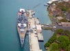 A large battleship is docked at a pier in a harbor, with adjacent buildings, roads, and greenery visible along the shoreline.