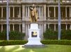 A bronze statue of a man in a cape and helmet stands on a white pedestal in front of a historic building with columns and arches.