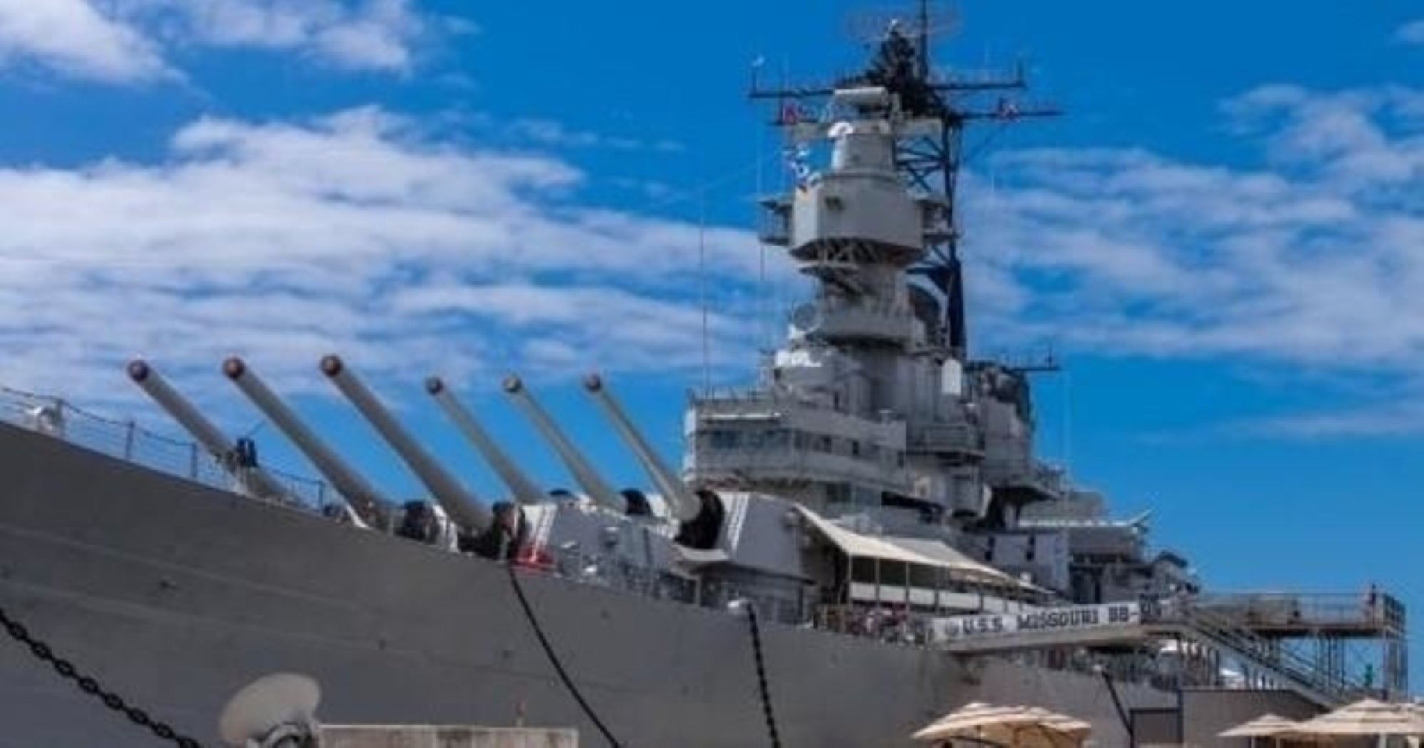 Gray battleship with large guns docked at a harbor under a partly cloudy blue sky; people are visible on deck near tents.
