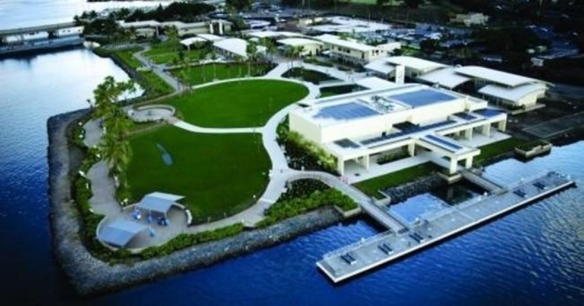 Aerial view of a waterfront park with green lawns, walking paths, covered seating areas, and a white building next to a dock, surrounded by water.