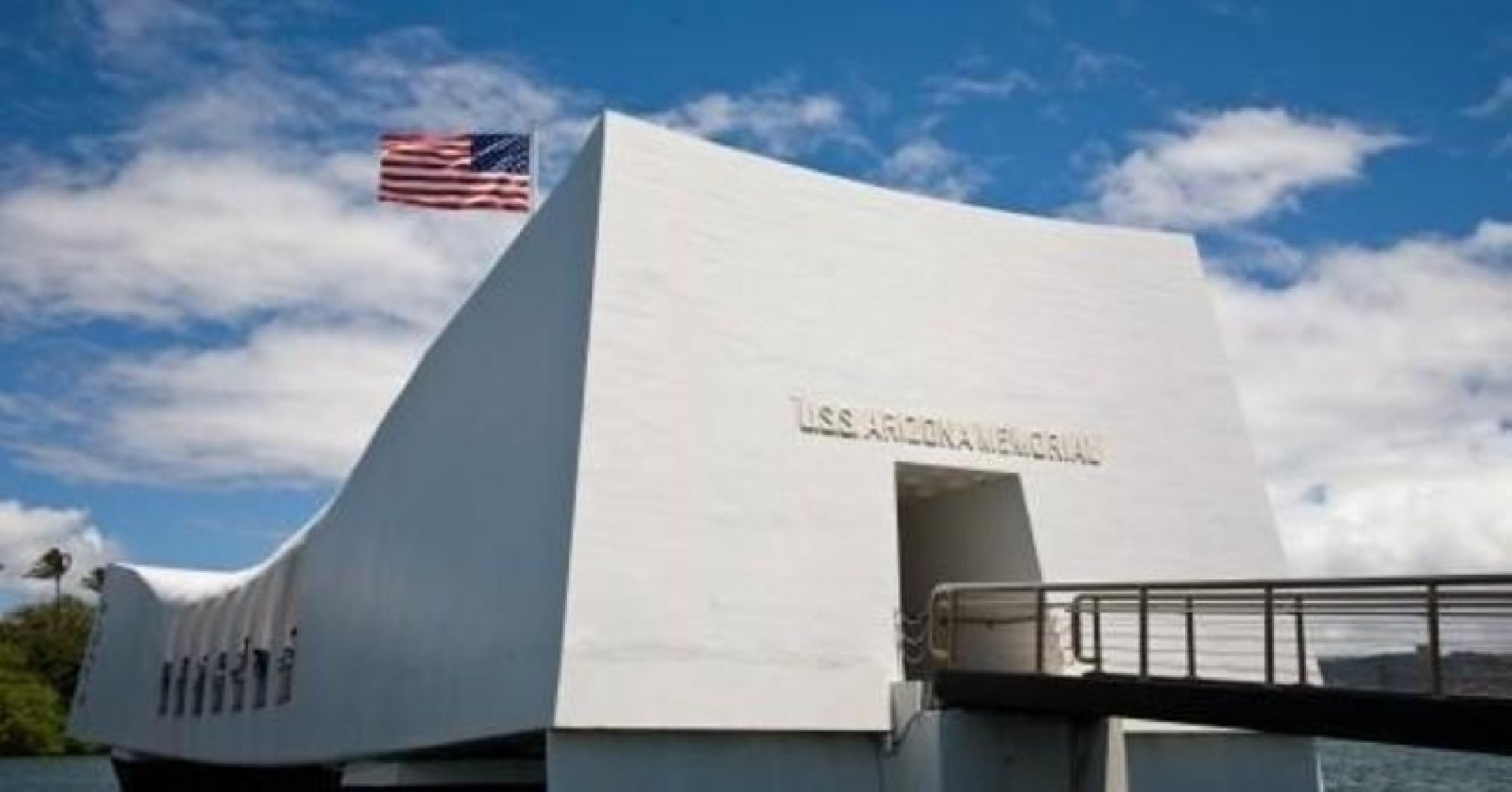 The USS Arizona Memorial, a white structure with an American flag, stands over the water at Pearl Harbor under a partly cloudy sky.