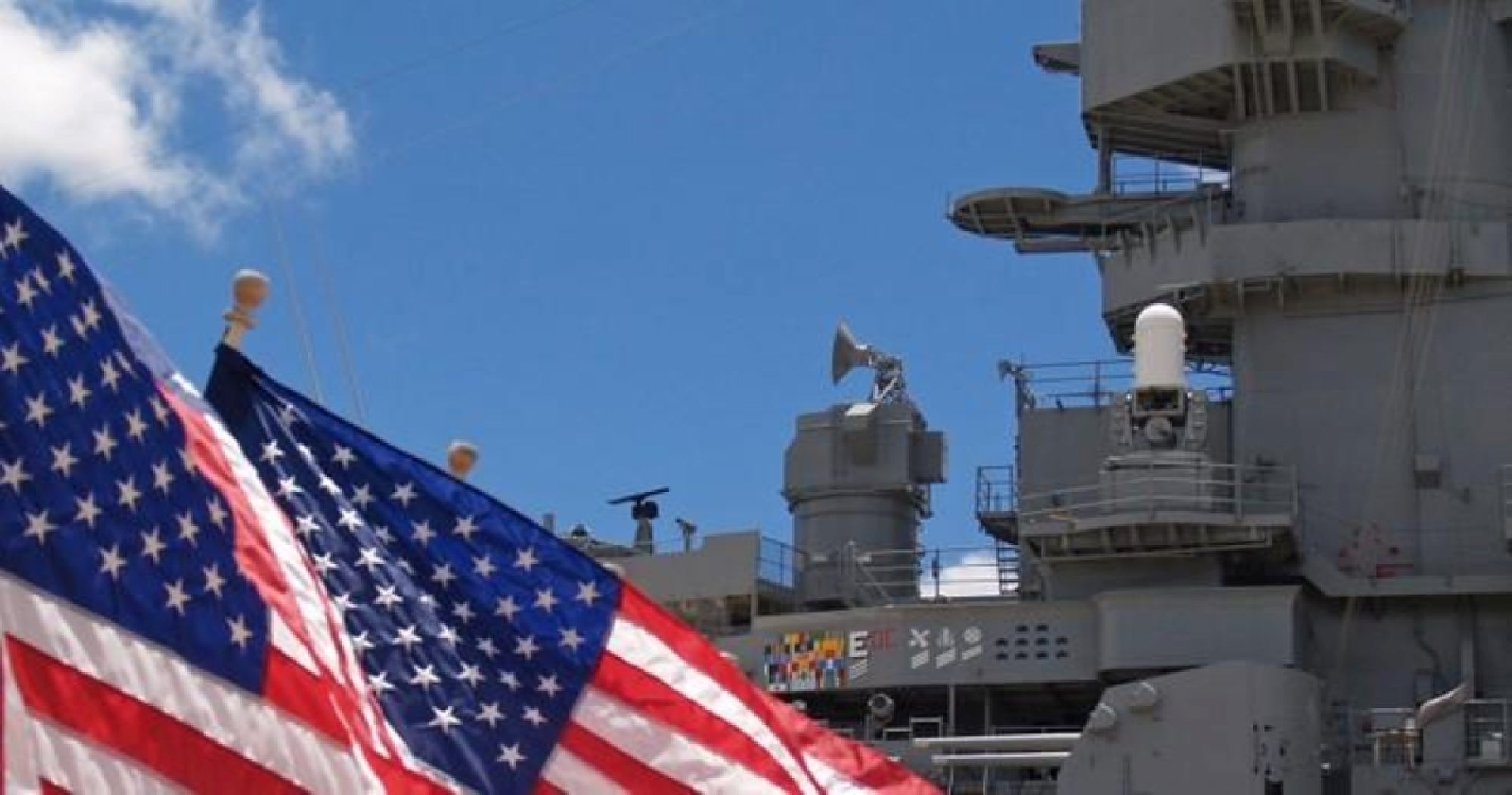 A close-up of an American flag in the foreground with part of a gray military ship and blue sky in the background.