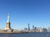 The Statue of Liberty stands on Liberty Island with the New York City skyline visible in the background under a clear blue sky.