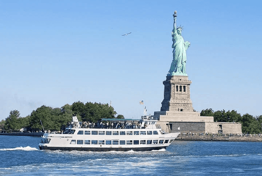 A ferry sails past the Statue of Liberty on a clear day, with the statue standing prominently on Liberty Island and the sky bright blue.