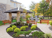 Outdoor patio with a covered gazebo, surrounded by benches and green shrubs. Two grills are positioned nearby, with a building and trees in the background.