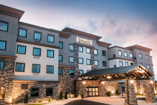 Front view of a modern hotel with a mix of stone and stucco facade, featuring a covered entrance and lit sign against a dusk sky.