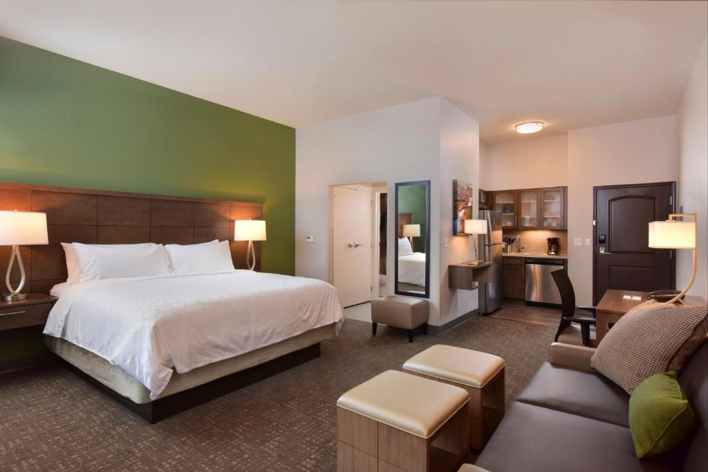 Hotel room with a king-size bed, green accent wall, two lamps, kitchenette, sofa, and a table with two chairs.
