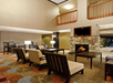 Hotel lobby with stone fireplace, multiple seating areas, and warm lighting. Carpeted floor with floral pattern and open space with a balcony above.