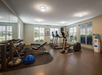 A gym room with treadmills, stationary bikes, weight racks, a bench, exercise balls, and a water cooler. Large windows provide natural lighting.