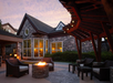 Stone house with large windows, outdoor patio seating, and a lit fire pit under a wooden canopy at sunset.