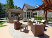 Outdoor patio area with wicker furniture, a stone fire pit, and a pergola. Brick building and trees in the background under a clear blue sky.