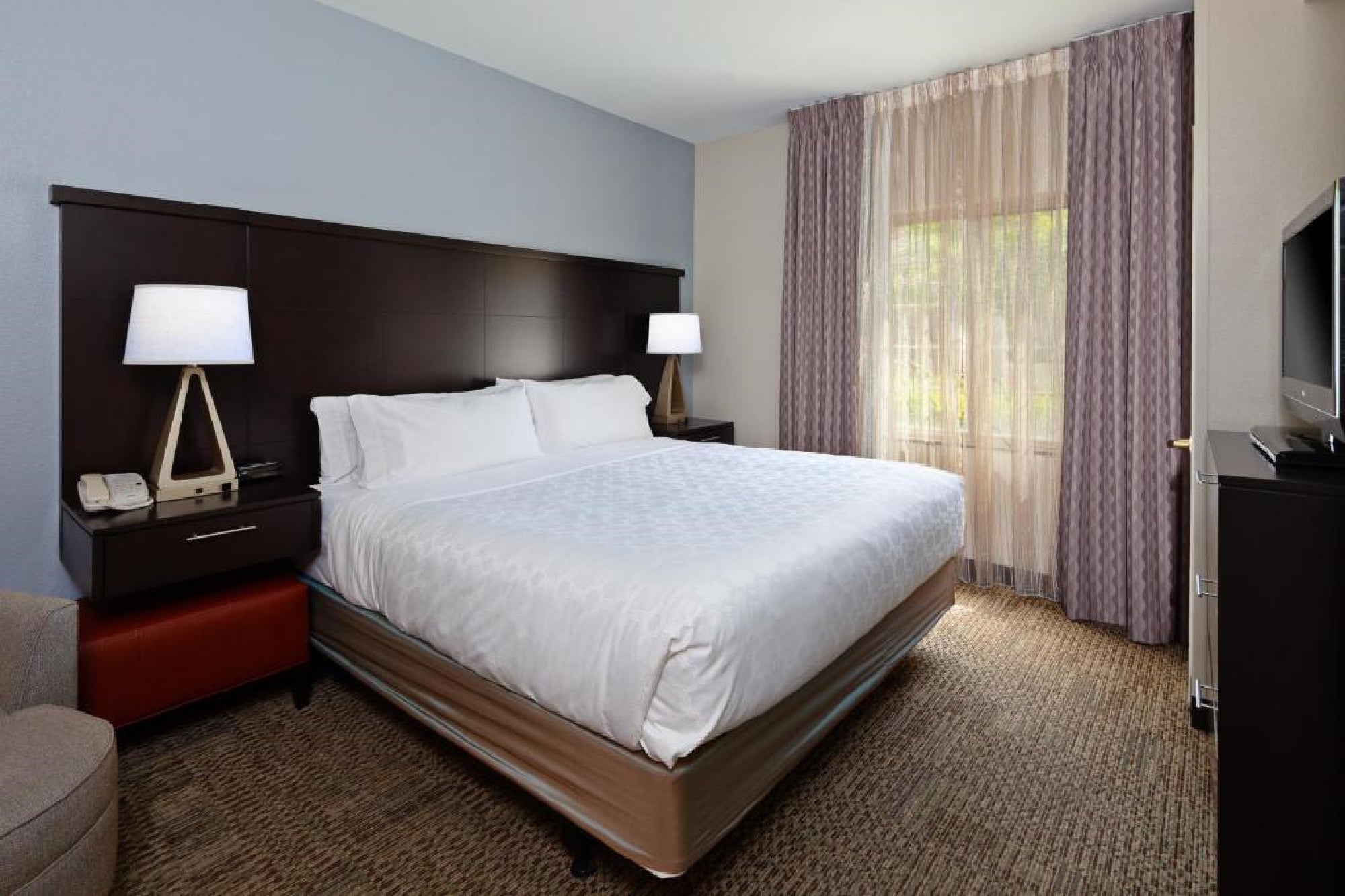 A hotel room with a large bed, two bedside lamps, an armchair, a window with curtains, and a TV.