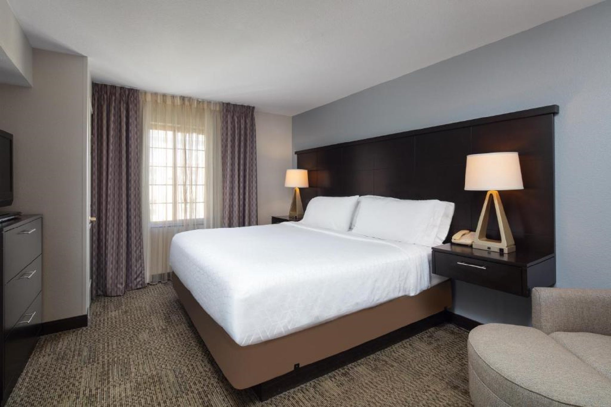 A hotel room with a king-size bed, two side tables with lamps, a window with curtains, a TV, dresser, and an armchair. The bedding is white, with neutral-toned walls and carpet.