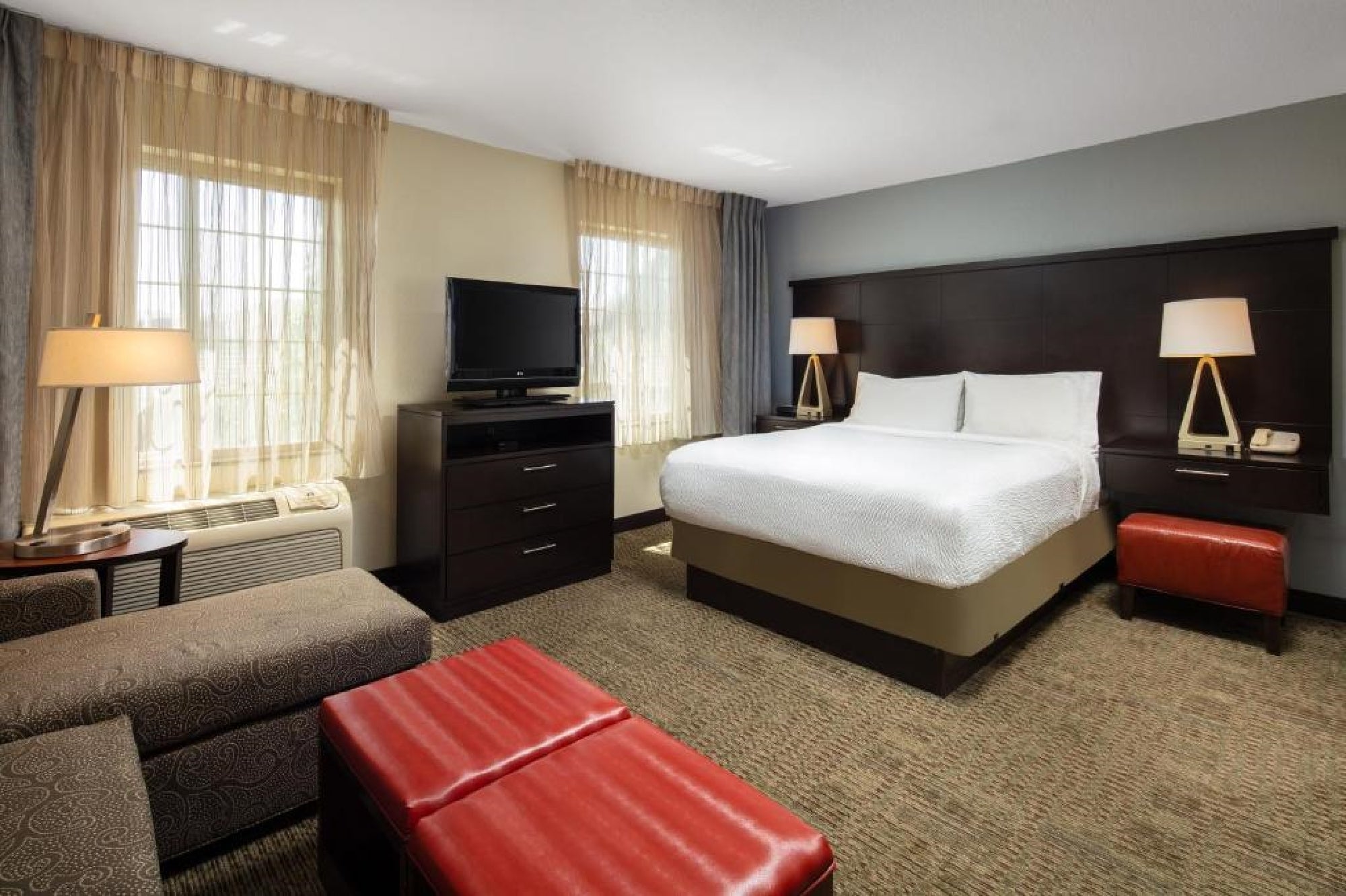 Hotel room with a double bed, two nightstands with lamps, a TV on a dresser, two red ottomans, and a patterned sofa. Natural light filters through two windows with sheer curtains.