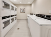 Laundry room with stacked dryers on the left and horizontal washers on the right, beige walls, tiled floor, and a framed picture on the wall.
