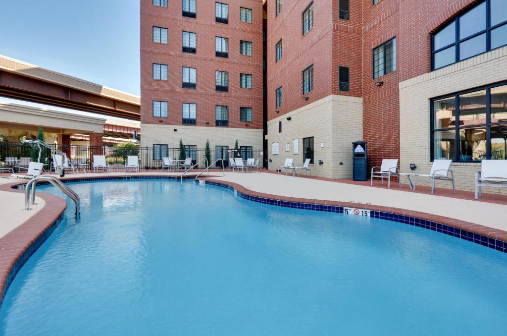 Outdoor pool surrounded by red brick buildings, with lounge chairs and a safety sign by the water. A partial view of a covered walkway is visible in the background.