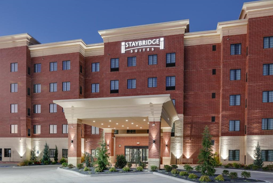 A multi-story brick hotel with a prominent "Staybridge Suites" sign, featuring large windows, an entrance canopy, and outdoor lighting. Landscaping includes small trees and shrubs.