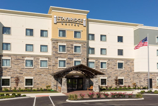 Exterior view of Staybridge Suites, a hotel building with a stone facade, multiple floors, and an American flag near the entrance.