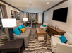 A modern living room with a gray sofa, blue and yellow cushions, two armchairs, a wooden coffee table, a large TV, and a dining area in the background. Carpet features a wavy pattern.