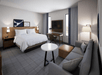 Modern hotel room with a king-sized bed, bedside tables, wall art, a work desk with chair, sofa, coffee table, floor lamp, and a flat-screen TV mounted on the wall.