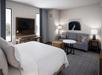 Modern hotel room with a king-size bed, sofa, TV, desk, and round coffee table. Decor includes a wall painting and floor lamp, with natural light from two windows.