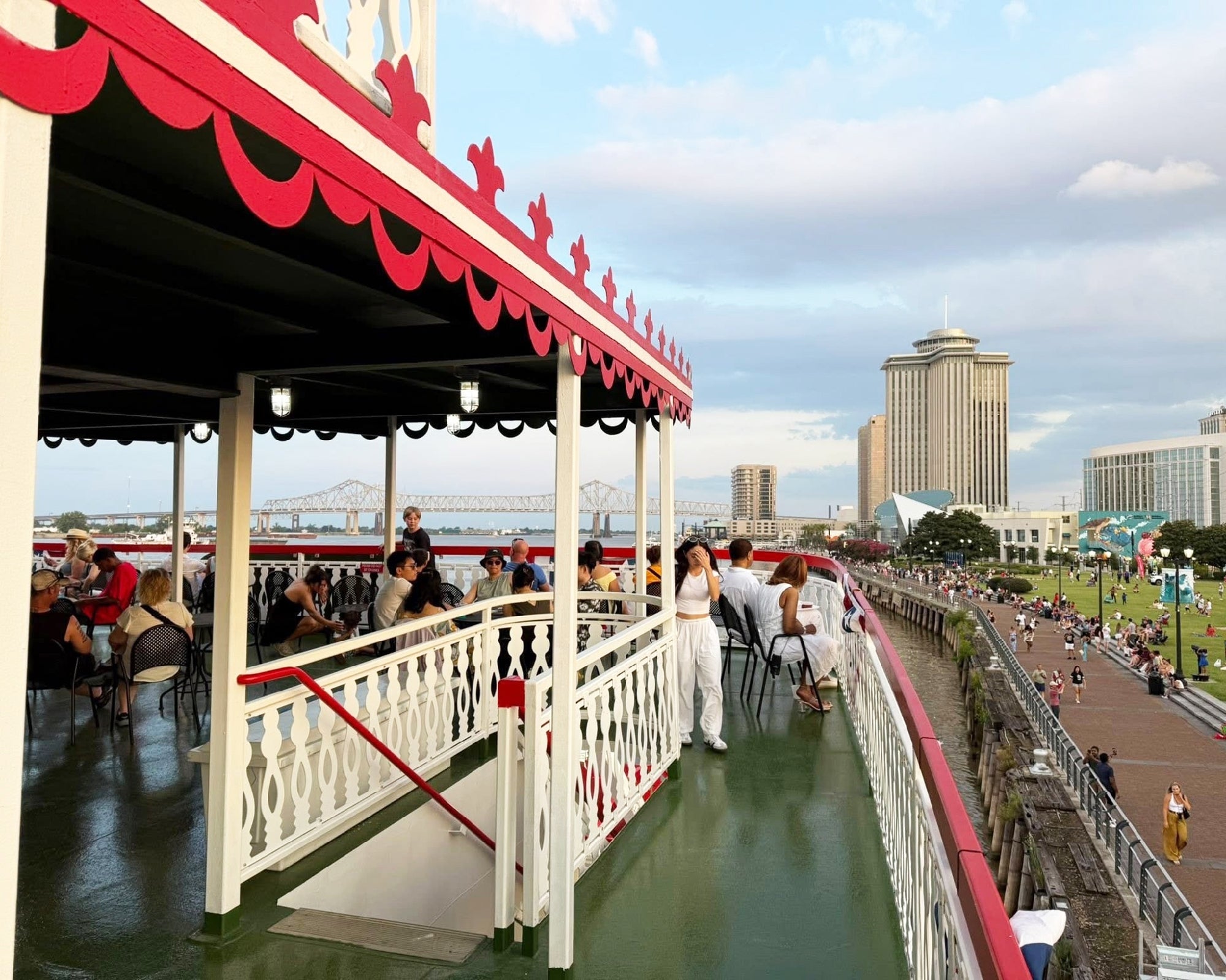 People sit and stand on the deck of a riverboat overlooking a waterfront park and cityscape, with tall buildings and a bridge visible in the background.