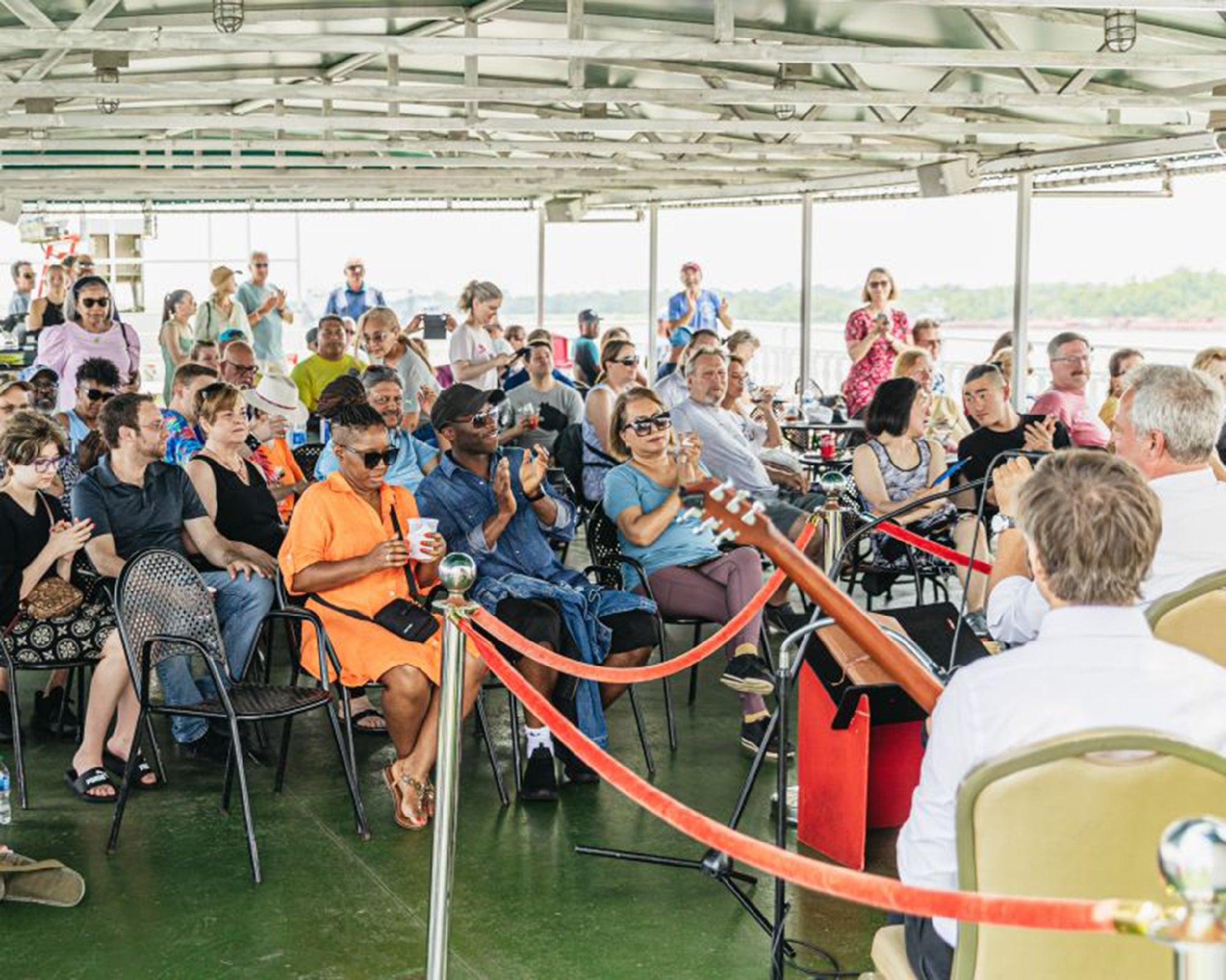 A group of people sit on a covered outdoor deck, watching a live musical performance. Some audience members are clapping, and musical instruments are visible in the foreground.