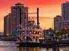 A steamboat docked at a riverside pier with tall buildings and a vibrant orange sky in the background during sunset.