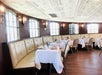 Elegant restaurant dining area with cream-colored tufted banquette seating, white tablecloths, and wooden floors, lit by natural light from large windows.