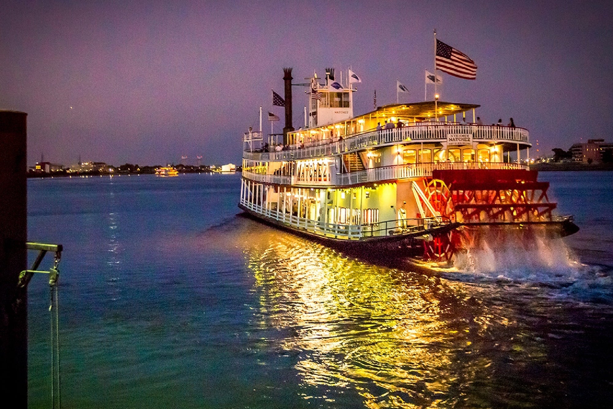 A brightly lit paddlewheel riverboat cruises on a calm river at dusk, with its reflection visible in the water.