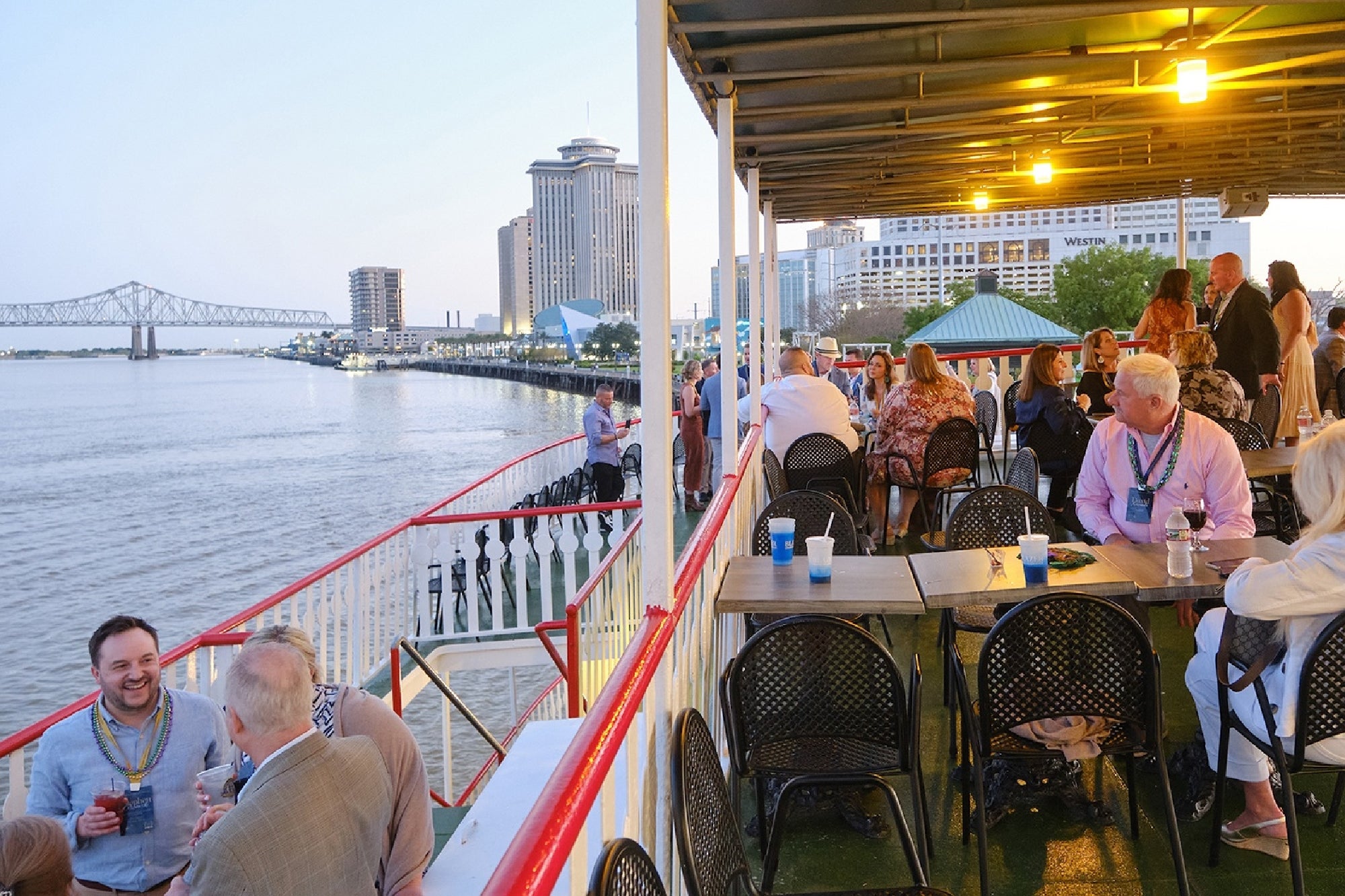 People sit and stand on the deck of a riverboat with drinks, overlooking a city skyline and a bridge at sunset.
