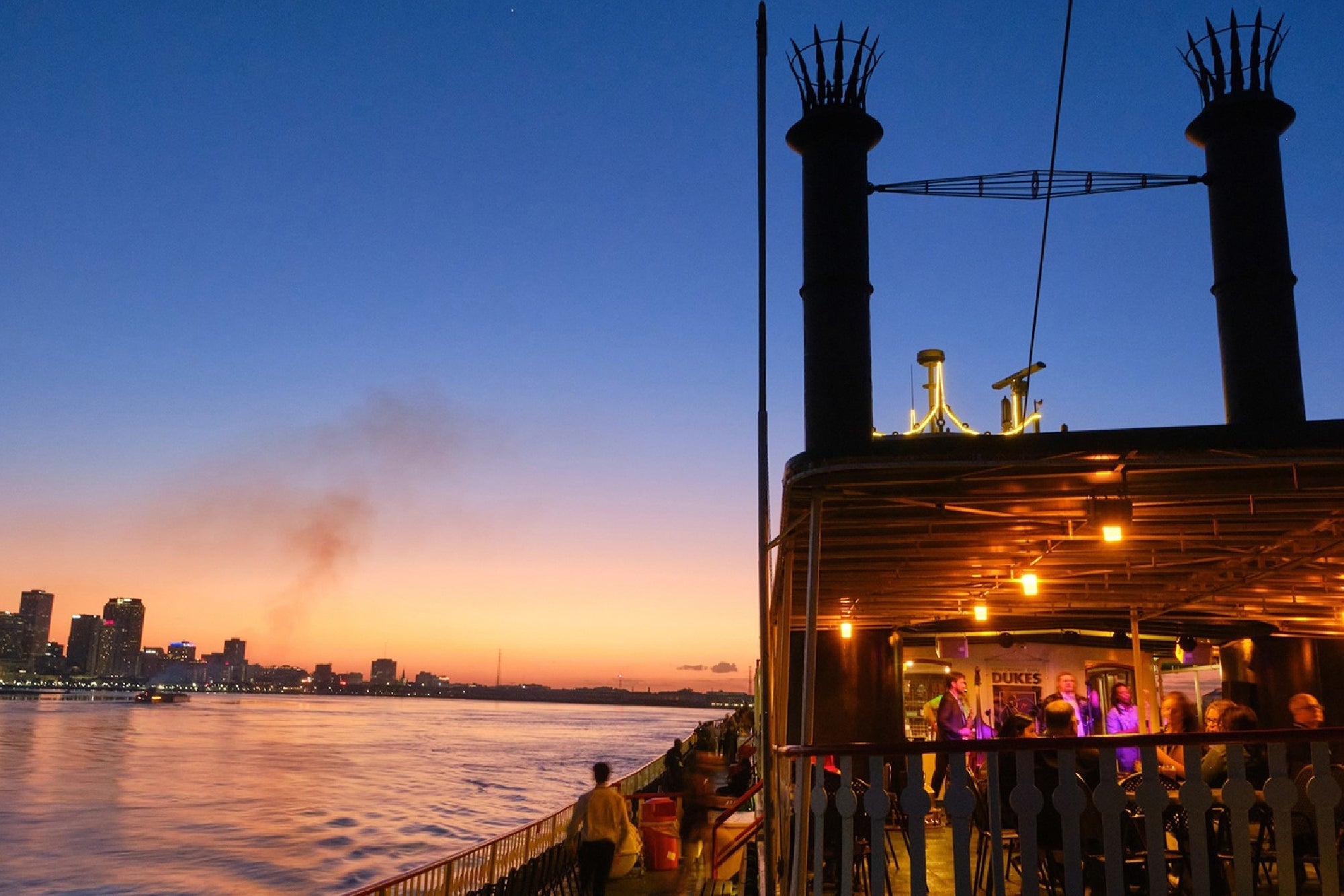 A paddle steamboat with illuminated lights travels on a river at sunset, with city buildings and smoke visible in the background.
