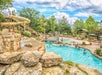 Outdoor water park with winding slide, rocks, trees, and a pool. People are swimming and lounging under umbrellas.