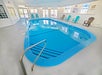 Indoor swimming pool with blue water, surrounded by several turquoise and brown chairs. White walls and windows in the background.