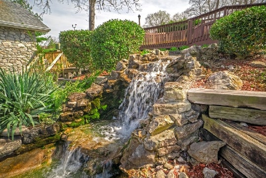A small waterfall cascades over rocks into a pond in a garden, surrounded by greenery and a wooden fence.