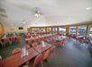 A spacious dining hall with multiple rows of tables and red chairs, ceiling fans, and large windows overlooking an outdoor area. Rolls of paper towels are placed on the tables.