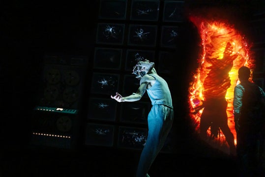 A person in light clothing poses dramatically in front of TV screens and an orange, glowing portal with shadowy figures visible inside.