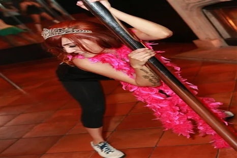 A woman with red hair wearing a bright pink feather boa and a crown while leaned over holding onto a silver stripper pole.