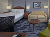 Hotel room with a bed, sofa, round table, and two lamps. A framed picture hangs on a light-colored wall.