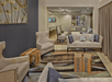 A modern lounge with beige sofas, armchairs, and decorative wood tables. Art pieces adorn the walls, and a TV is mounted in the background. The room is softly lit and carpeted.