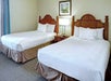 Two double beds with white bedding in a hotel room, wooden headboards, a bedside table with a lamp, and a framed picture on the wall.