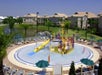 Colorful water playground with slides and sprinklers in a circular pool, surrounded by lounge chairs and umbrellas, near residential buildings and palm trees under a clear blue sky.