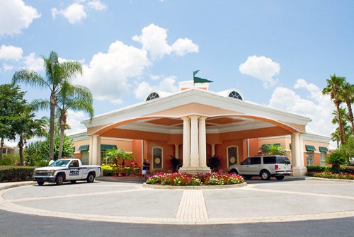 Entrance of a building with a circular driveway, two parked vehicles, and a flower bed in front. The building has a light orange exterior with four columns and a green flag on the roof.